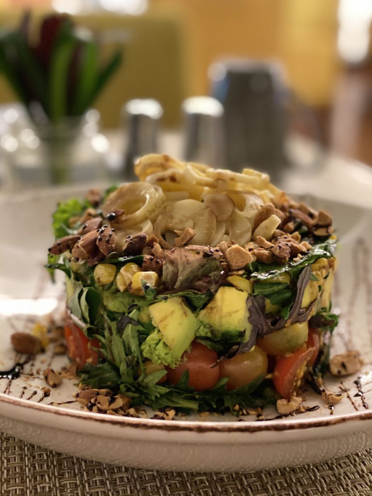 Layered salad with cherry tomatoes, avocado, greens, corn, hearts of palm, and crushed almonds